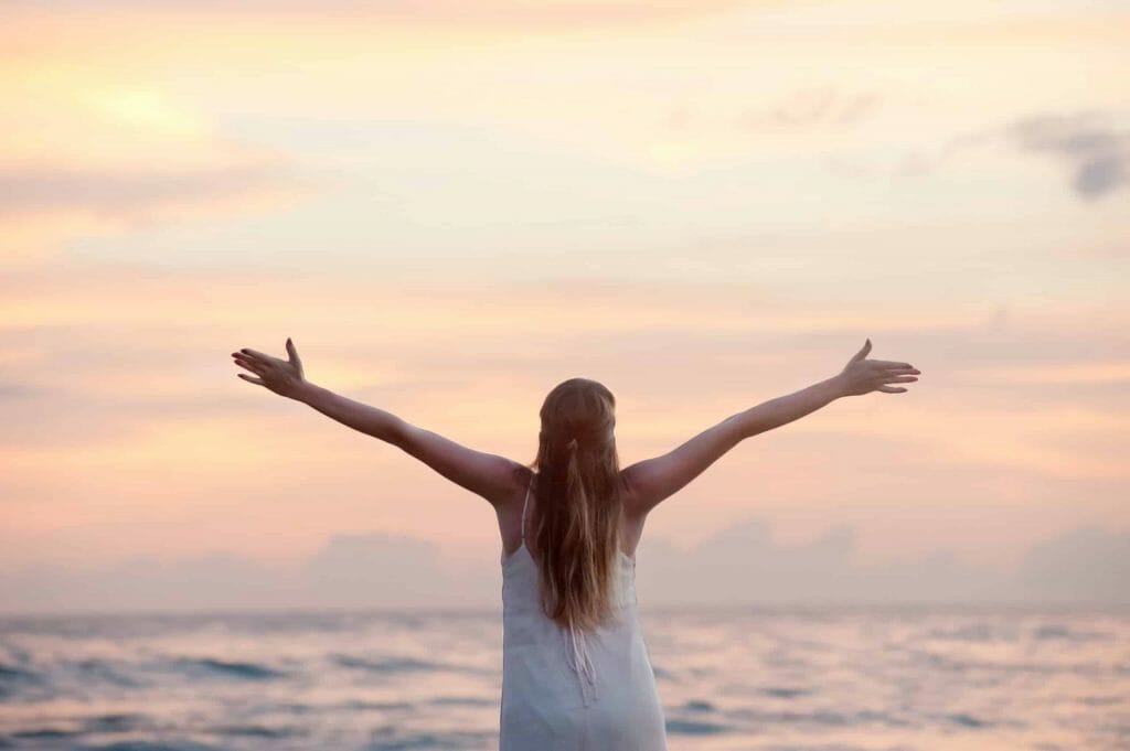 A woman facing away from the camera in front of the ocean with her arms outstretched